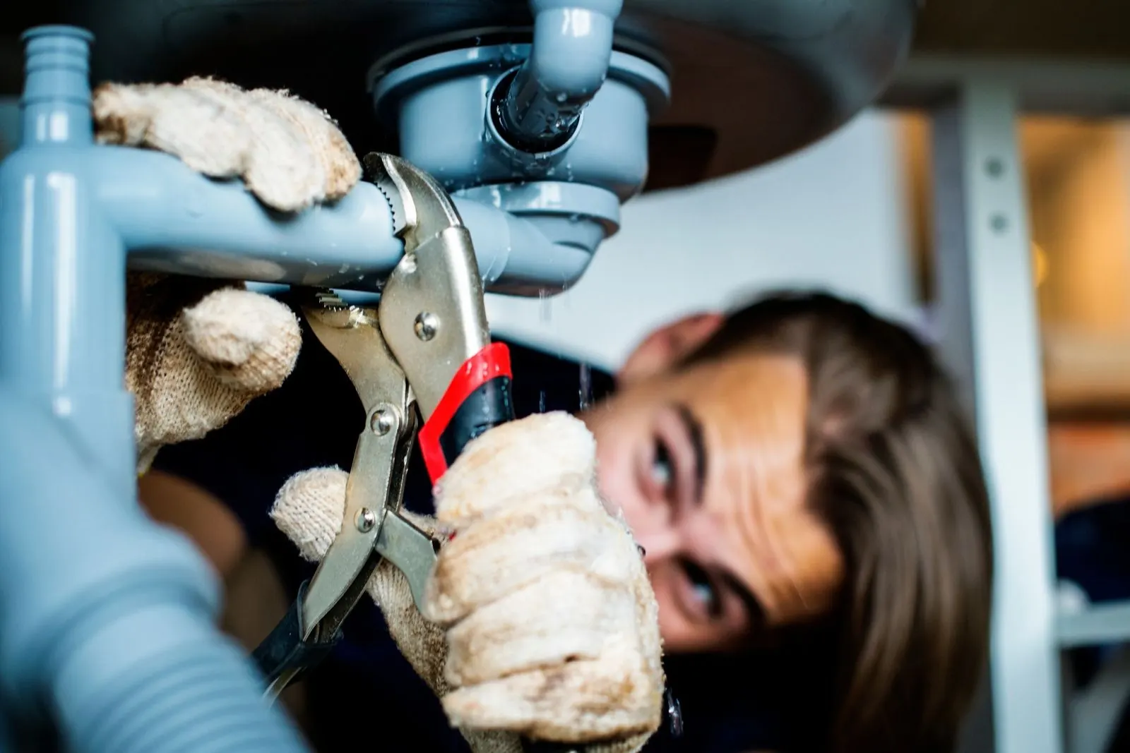 Plumber in gloves gripping a pipe fitting under a sink during an emergency repair